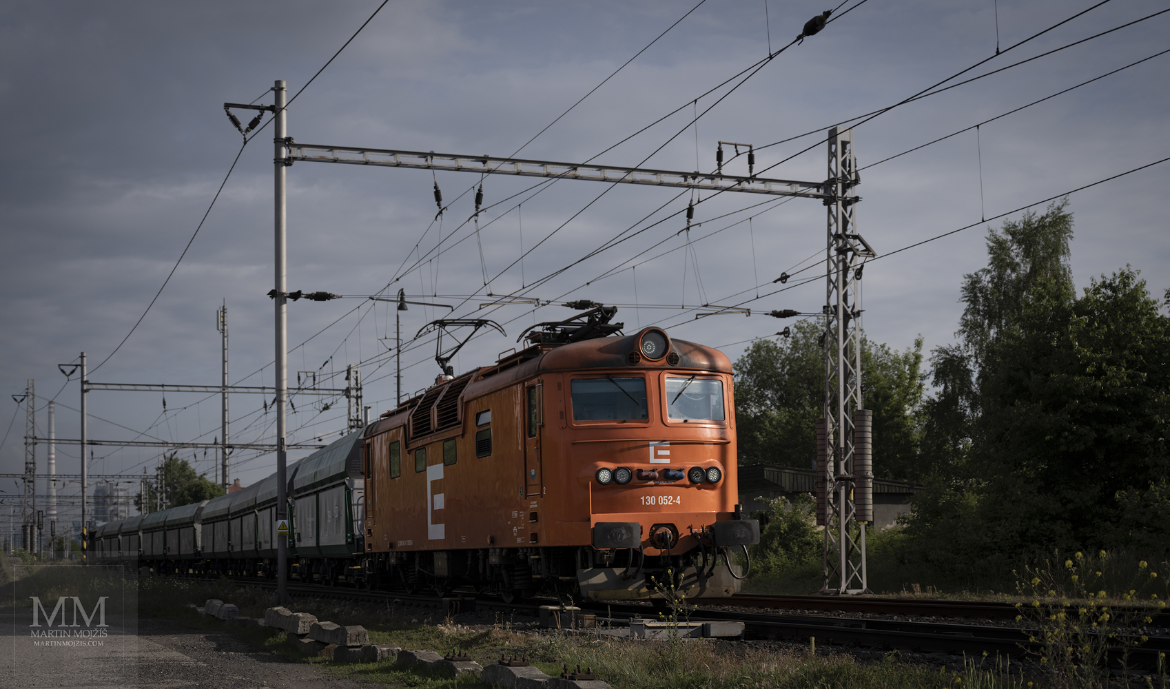 Oranžová lokomotiva 130 052-4 vyjíždí ze stanice v čele nákladního vlaku ve směru Kralupy nad Vltavou.