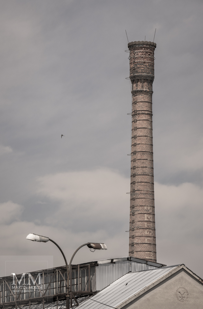 Vysoký cihlový tovární komín nad střechami továrny. Umělecká fotografie s názvem U TOVÁRNY II. Fotograf Martin Mojžíš.