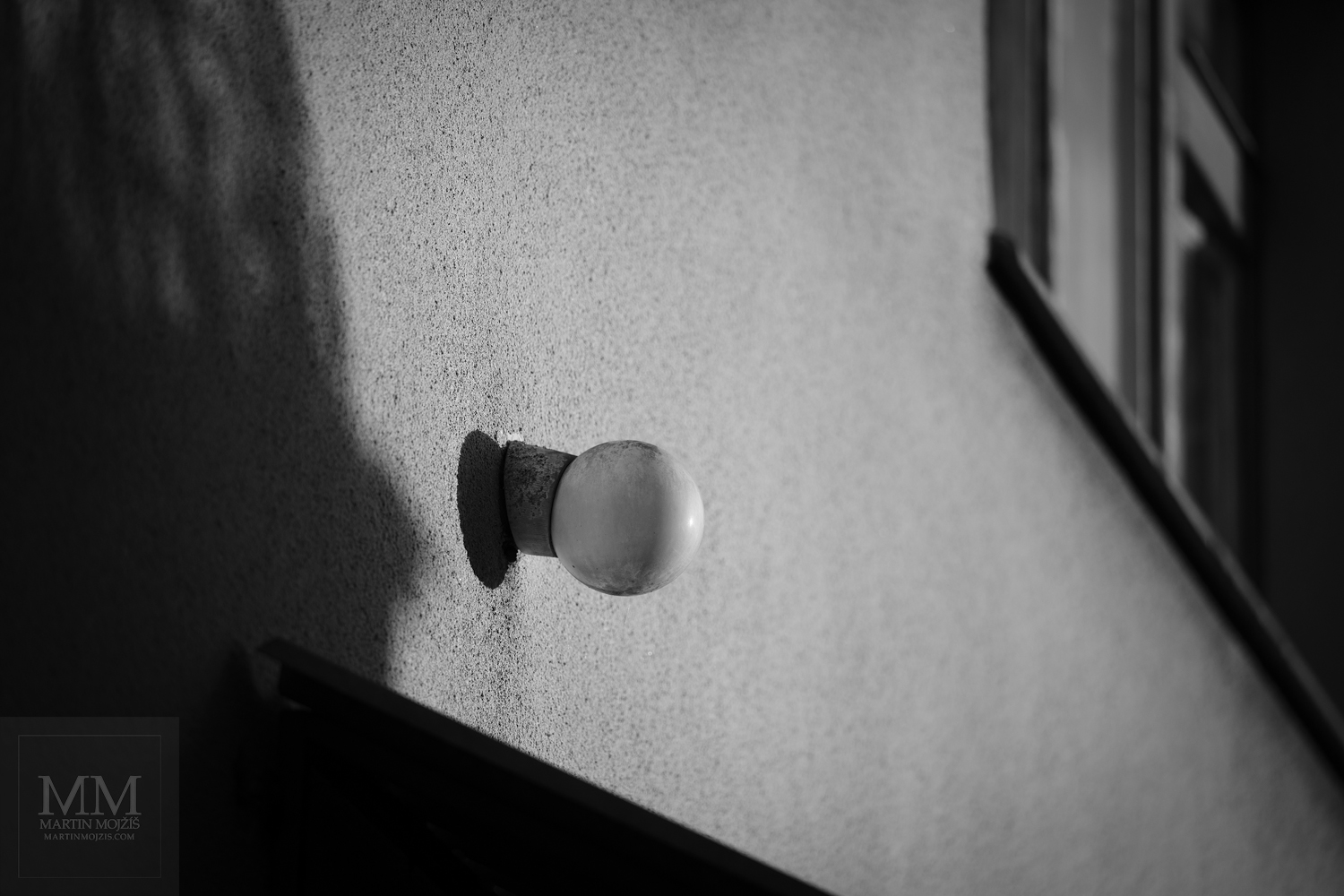 A lamp on a wall of a house in the light of the sun's rays. Fine art black and white photograph LAMP AND SUN, photographed by Martin Mojzis.