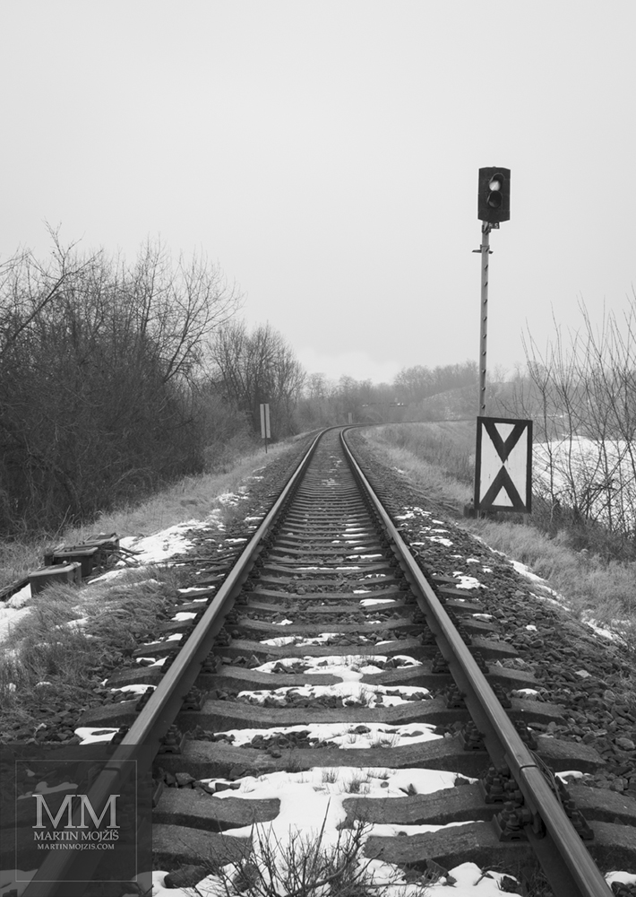 Jednokolejná železniční trať v zimě, vpravo světelné návěstidlo (semafor). Umělecká černobílá fotografie Martina Mojžíše s názvem NA POČÁTKU CEST.