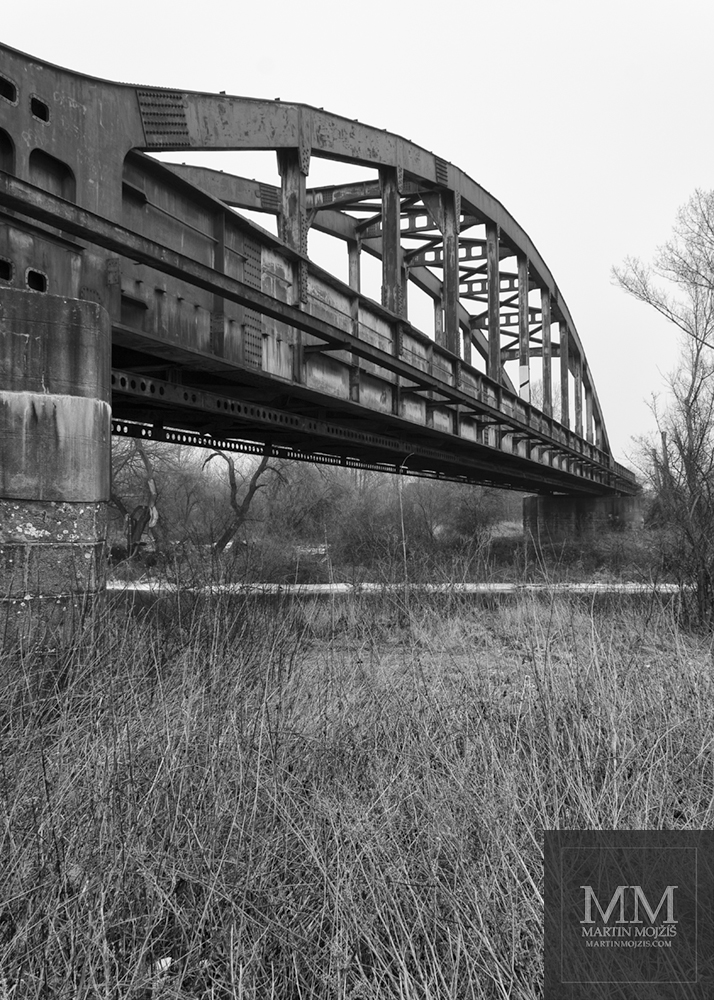 Černobílá umělecká fotografie ocelového železničního mostu s názvem NAD ŘEKOU. Fotograf Martin Mojžíš.